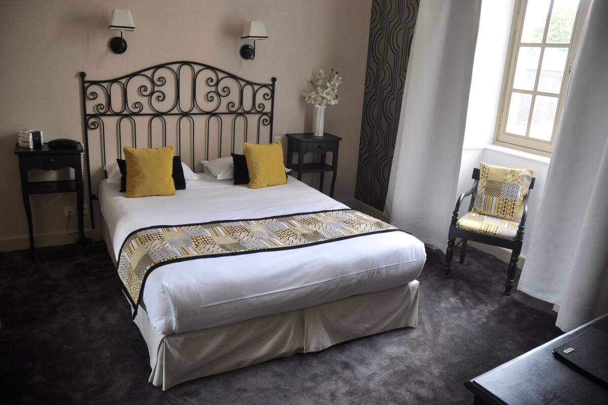 Un lit bien fait, recouvert d’une couvre-lit à motifs, deux lampes de chevet, une table de chevet avec un téléphone, une chaise agrémentée d’un coussin à motifs et une fenêtre avec des rideaux.