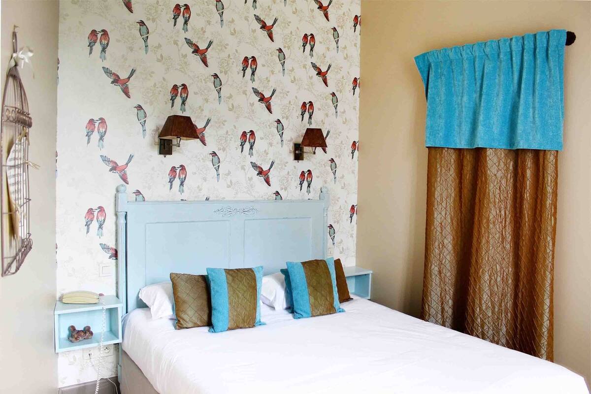 Une chambre à coucher avec un lit, des oreillers, une table de chevet, une lampe, un téléphone et un mur décoré d’un motif d’oiseaux.