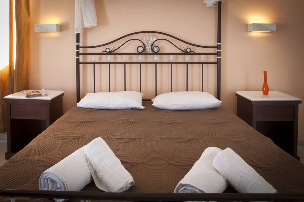 A neatly made bed with white pillows and towels, a metal headboard, and bedside tables with a vase on one.