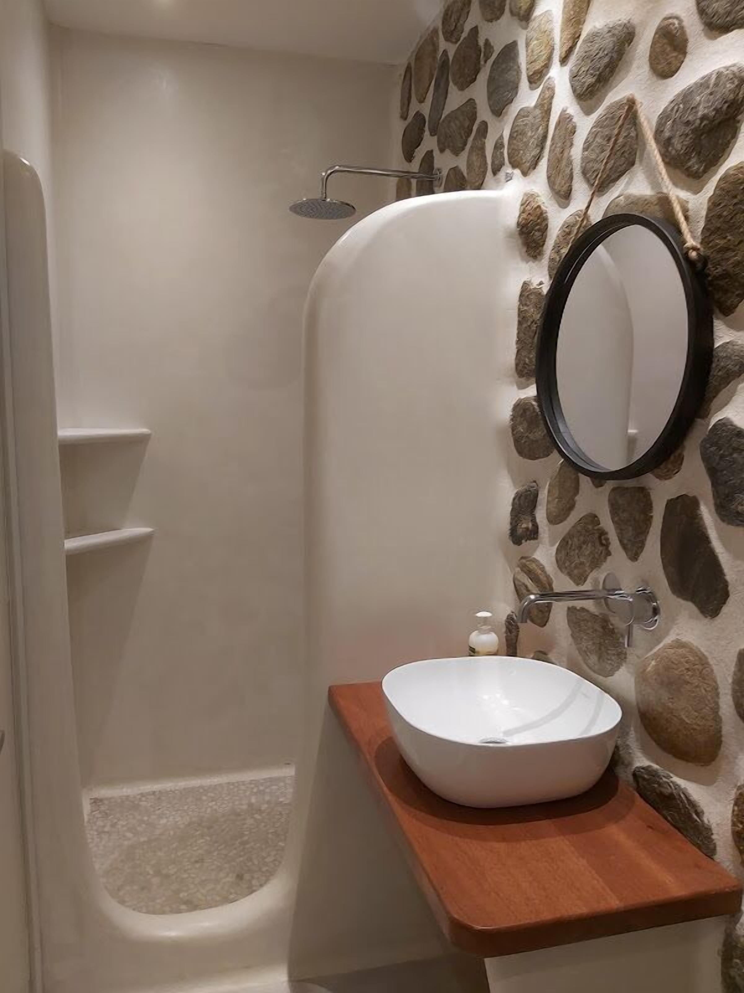 A bathroom with a stone wall, a bathtub, a round mirror, and a wooden vanity with a sink.