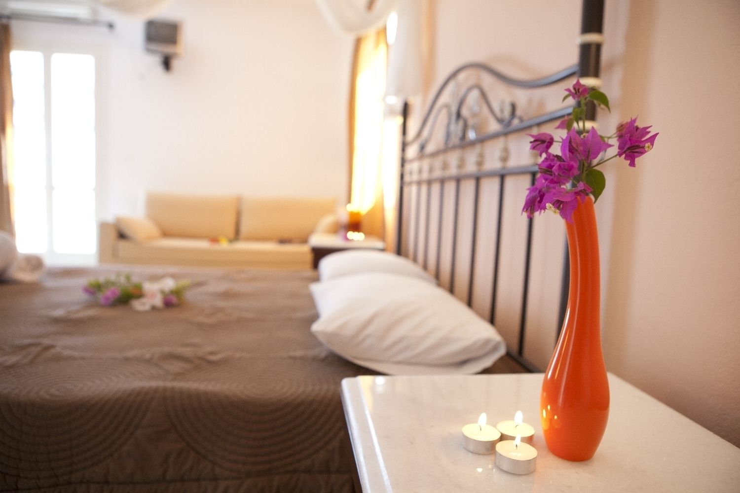 Una habitació d'hotel amb un llit, una tauleta de nit amb espelmes i un vas amb flors.