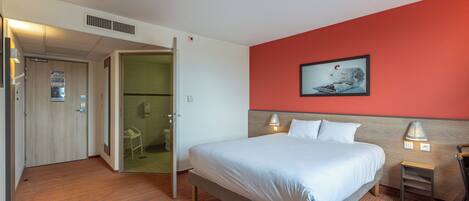 Una habitación de hotel con cama, una silla, un escritorio y un baño visible a través de una puerta abierta.
