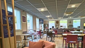 Een moderne hotellobby met een zithoek, houten tafels en stoelen. Grote ramen zorgen voor natuurlijk licht en uitzicht op de omgeving.