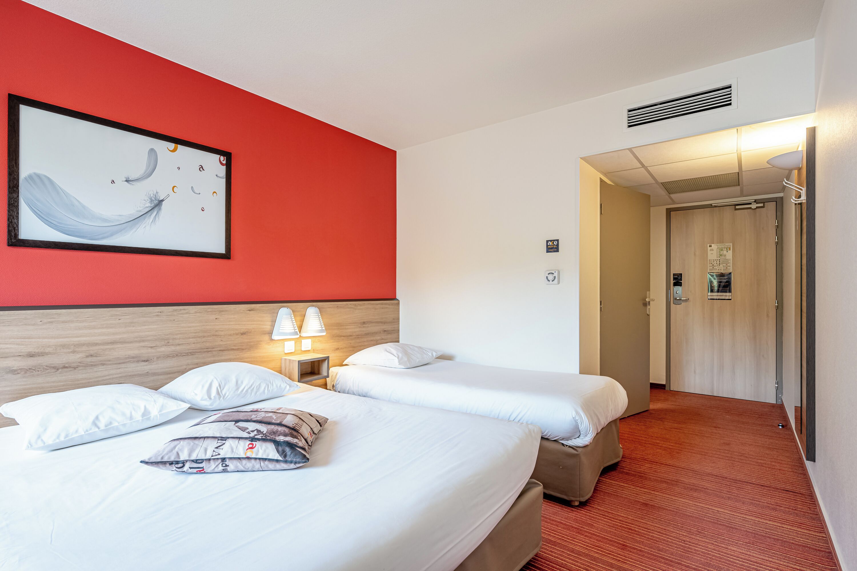 Quarto de hotel com duas camas, uma parede de destaque vermelha, uma obra de arte emoldurada e um corredor que leva a uma porta.
