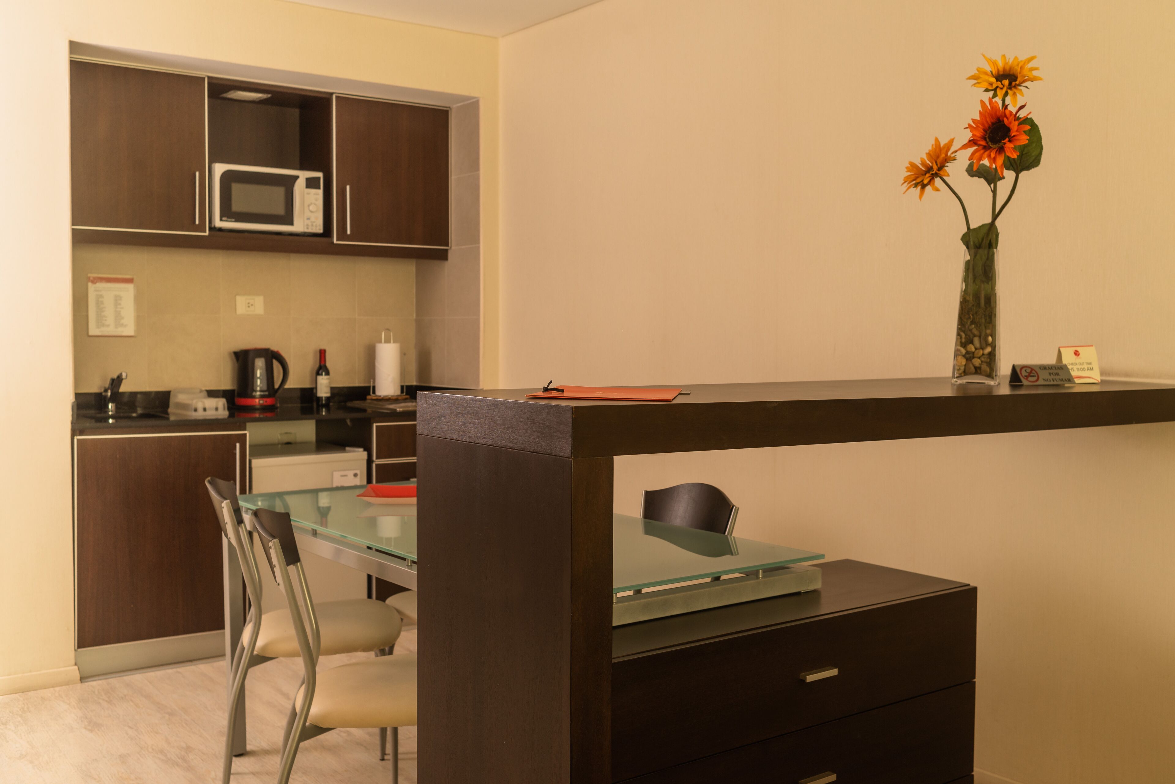 A modern kitchen with dark wooden cabinets, a microwave, and a glass table with a chair. There is a vase with flowers on the table.