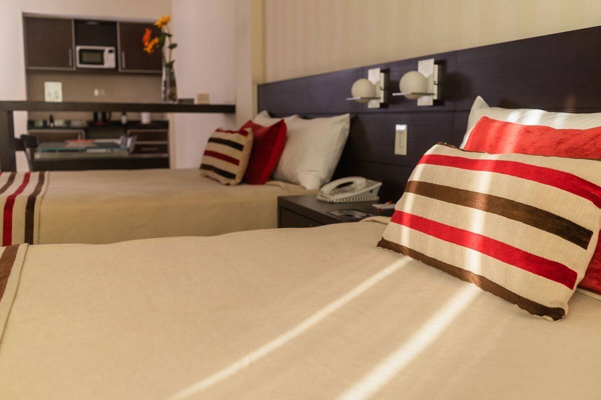 A hotel room with two beds, a bedside table with a phone, a small desk with a lamp, and a kitchenette in the background.
