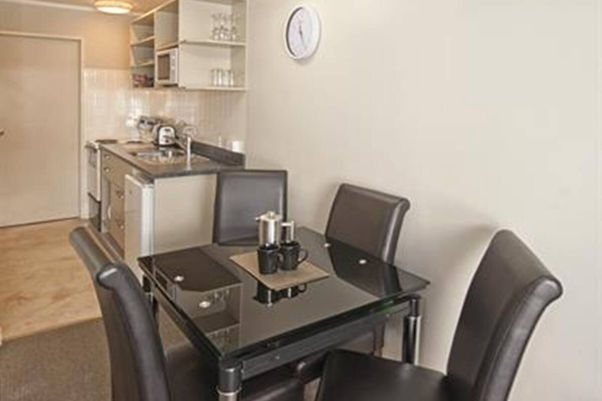 A compact dining area with a glass table and four chairs, a kitchen in the background, and a clock on the wall.
