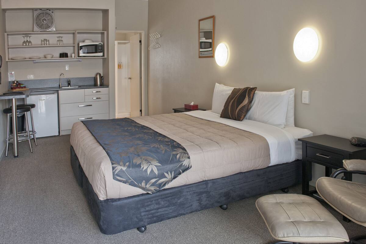 A compact hotel room with a bed, a desk with a chair, a microwave, and a small kitchenette.