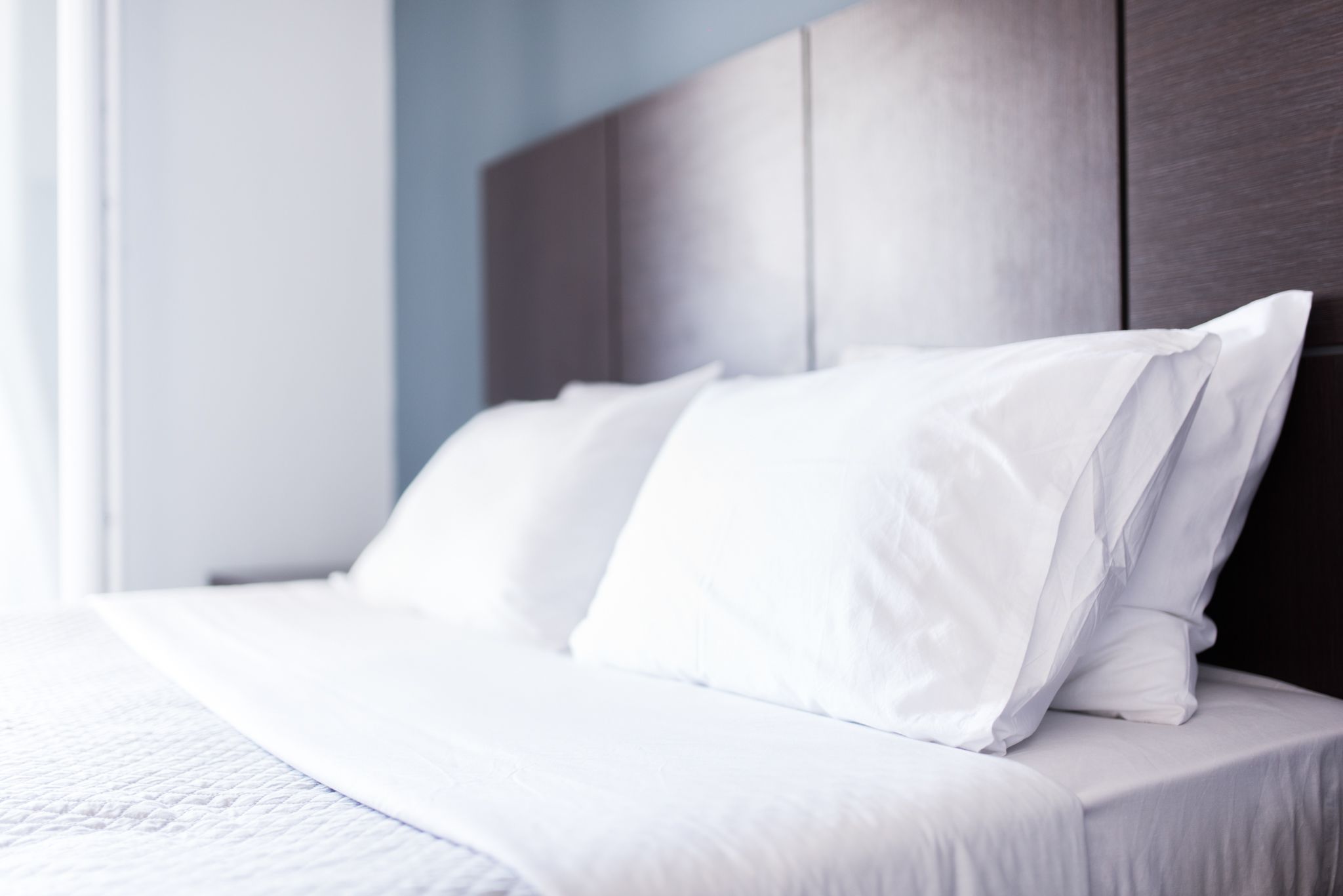 Una cama bien hecha con sábanas y almohadas blancas, un cabecero de madera y una pared de color claro.