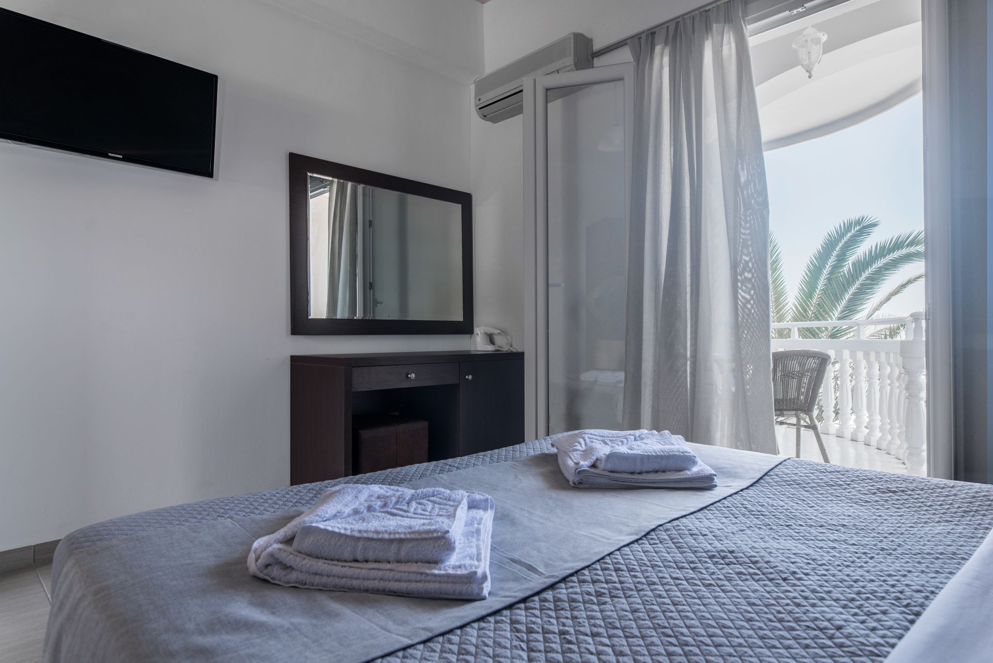A hotel room with a bed, a TV mounted on the wall, a small desk, and a balcony with a chair.