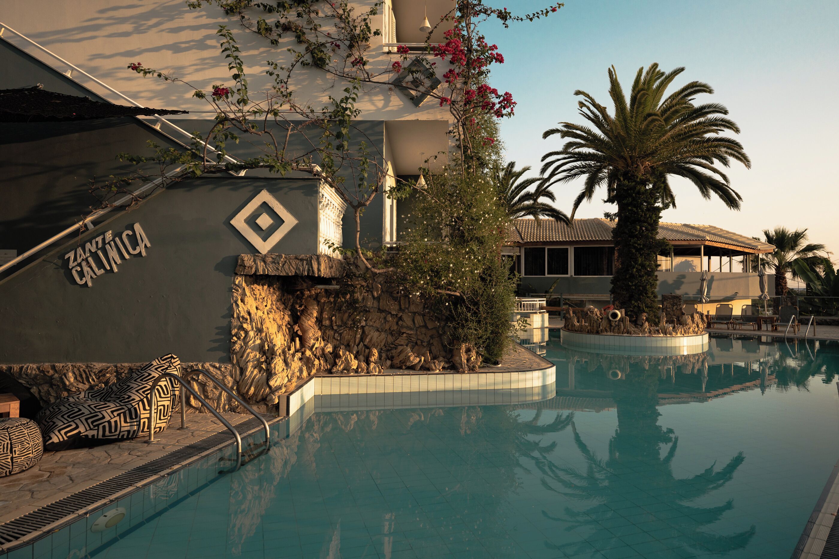 A swimming pool with a stone wall, palm trees, and a building with a sign reading "ZANTE CALINICA".