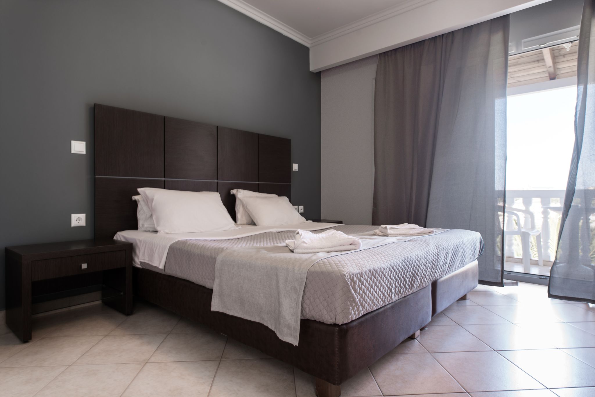 Une chambre à coucher moderne avec un grand lit, une tête de lit sombre et une vue sur l’extérieur grâce à un balcon.