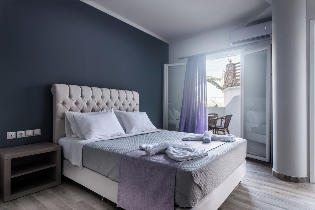 A modern bedroom with a tufted headboard, a neatly made bed with white linens, a small bedside table, and a view of the outside through a sliding glass door.