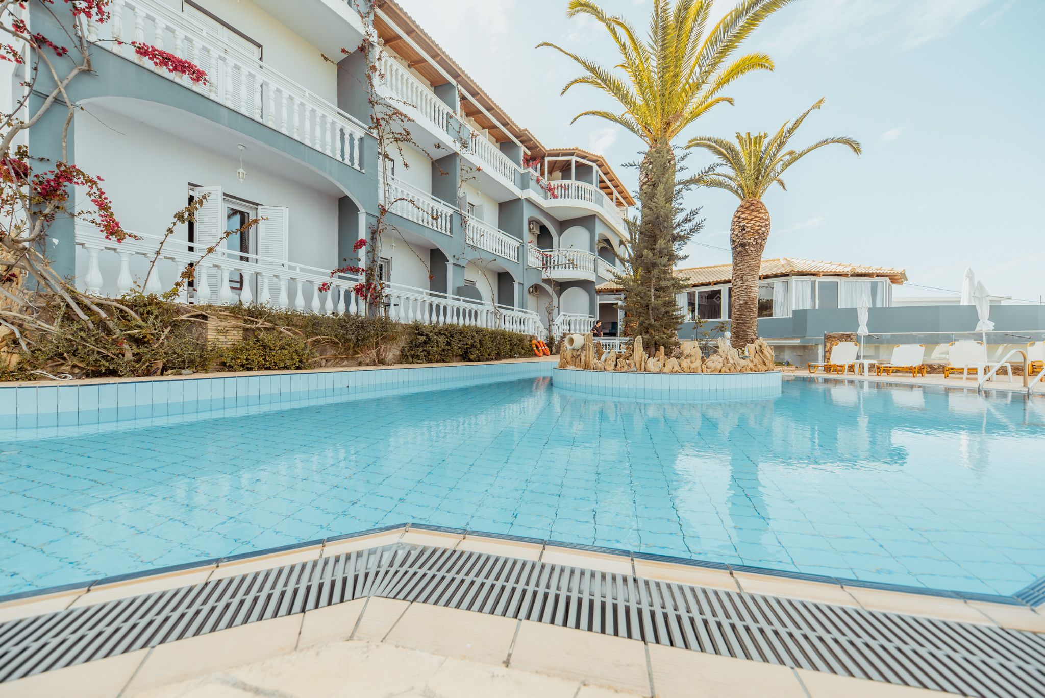 A swimming pool with a tiled deck, surrounded by a building with balconies and a palm tree.