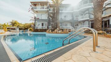 A swimming pool with a tiled deck, surrounded by palm trees and a multi-story building with balconies.
