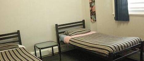 A small, simple room with two single beds, a small table, and a window with curtains.