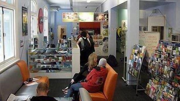 A library with bookshelves, a reading area with chairs, and a counter with a person behind it.