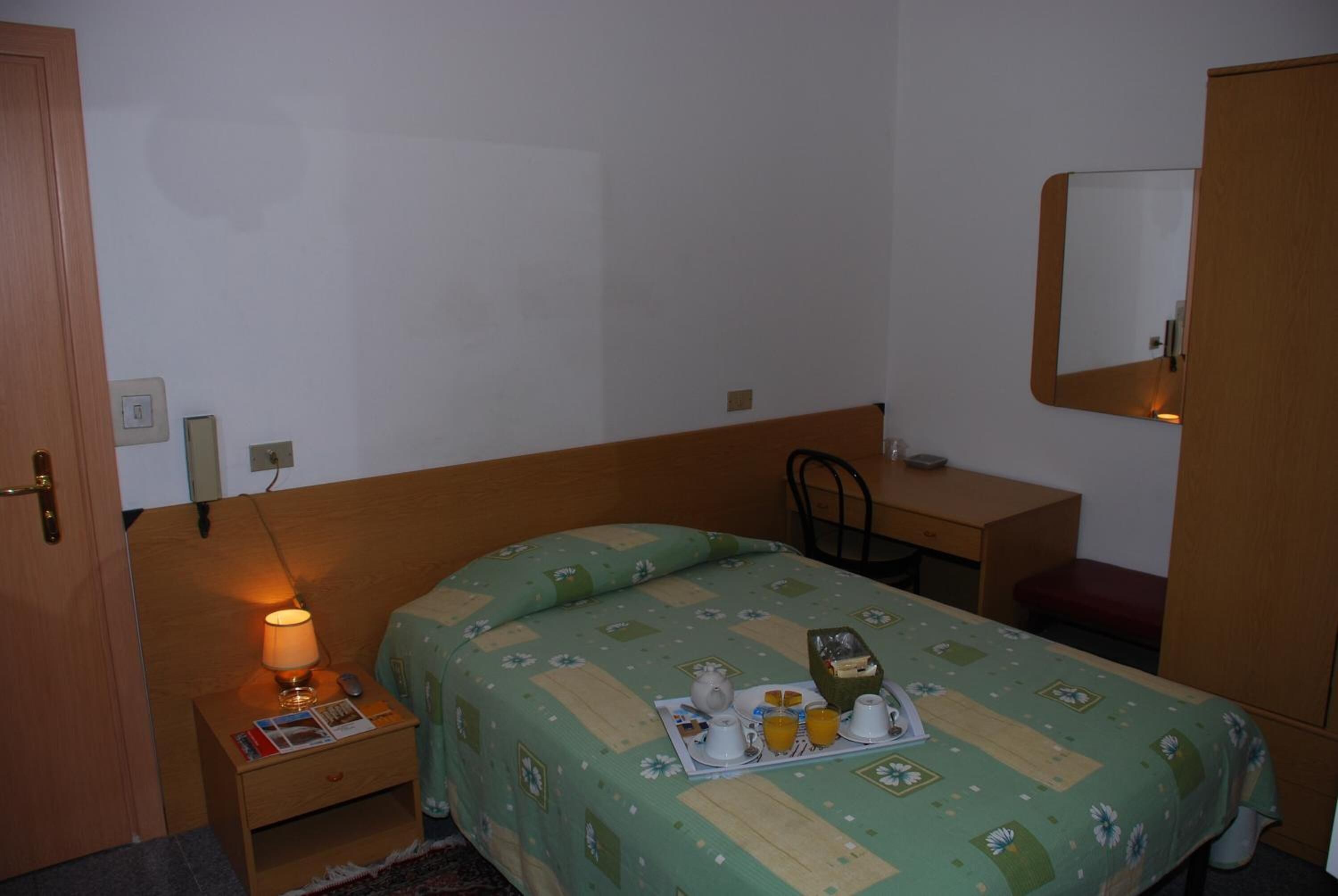 A small, simple room with a bed, a nightstand with a lamp, a desk, and a mirror.