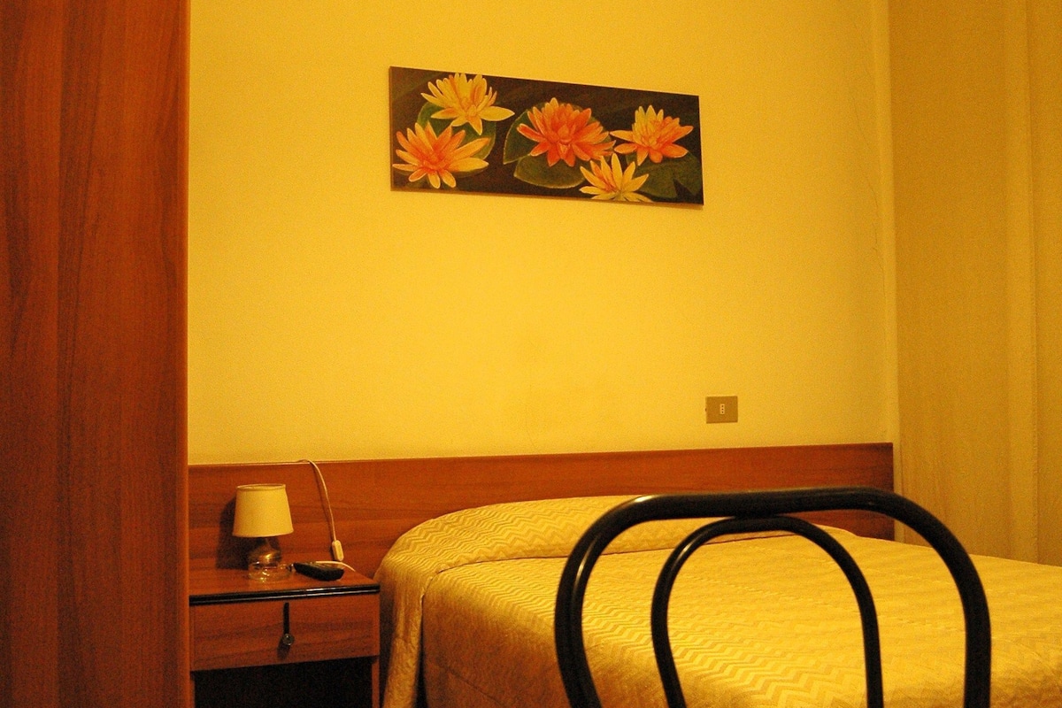 A hotel room with a bed, a wooden nightstand, a lamp, and a painting of flowers on the wall.