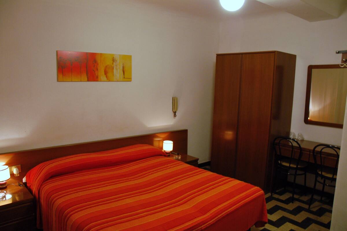 A hotel room with a bed, a desk, a wardrobe, and a painting on the wall.