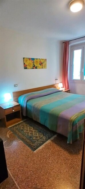 Una camera da letto con un letto, un comodino, una lampada, una finestra con tende e un quadro appeso al muro.