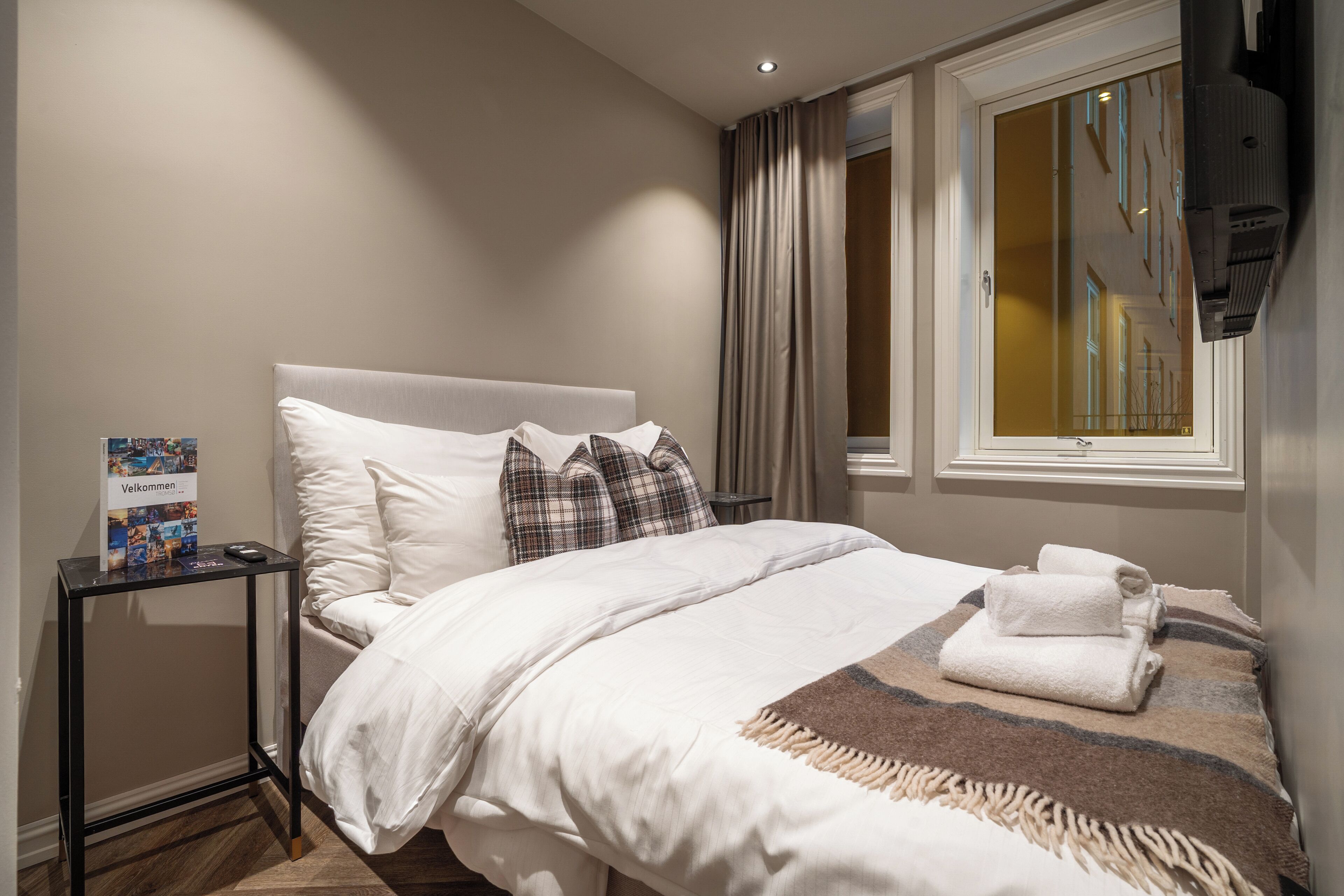 A hotel room with a bed, a bedside table, a window with curtains, and a view of the building outside.