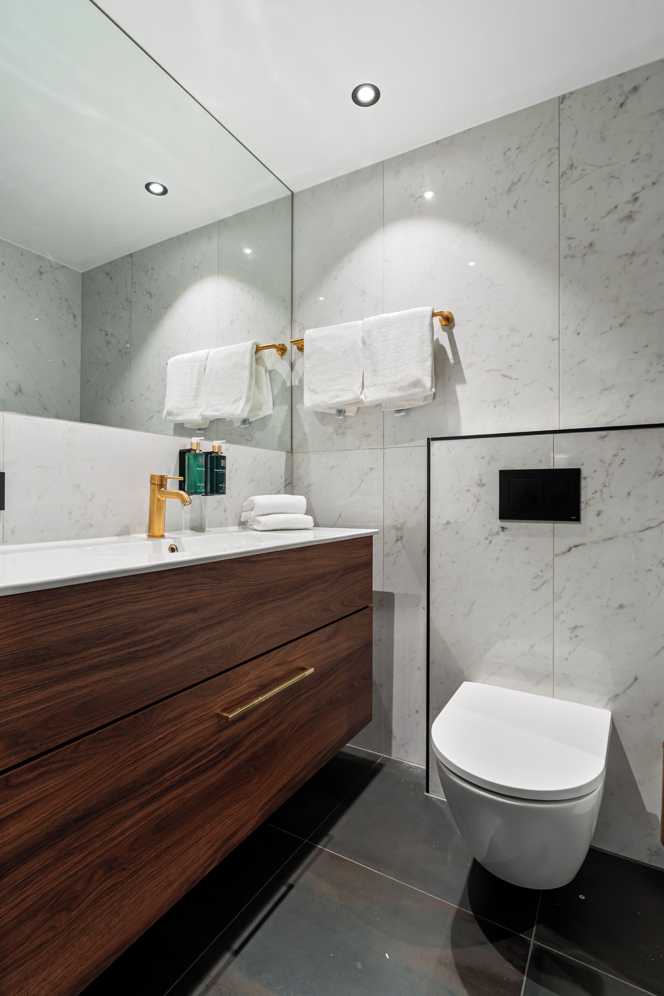 A modern bathroom with a marble wall, a wooden vanity, a white toilet, and a large mirror.