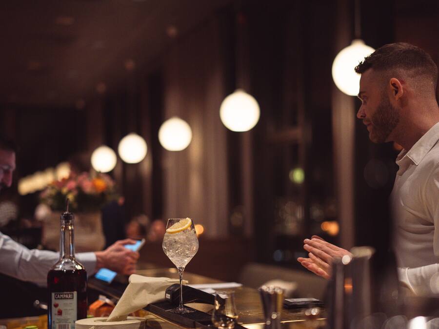 A bar setting with a bottle of Campari, a glass with ice and a lemon slice, and a person in a white shirt.
