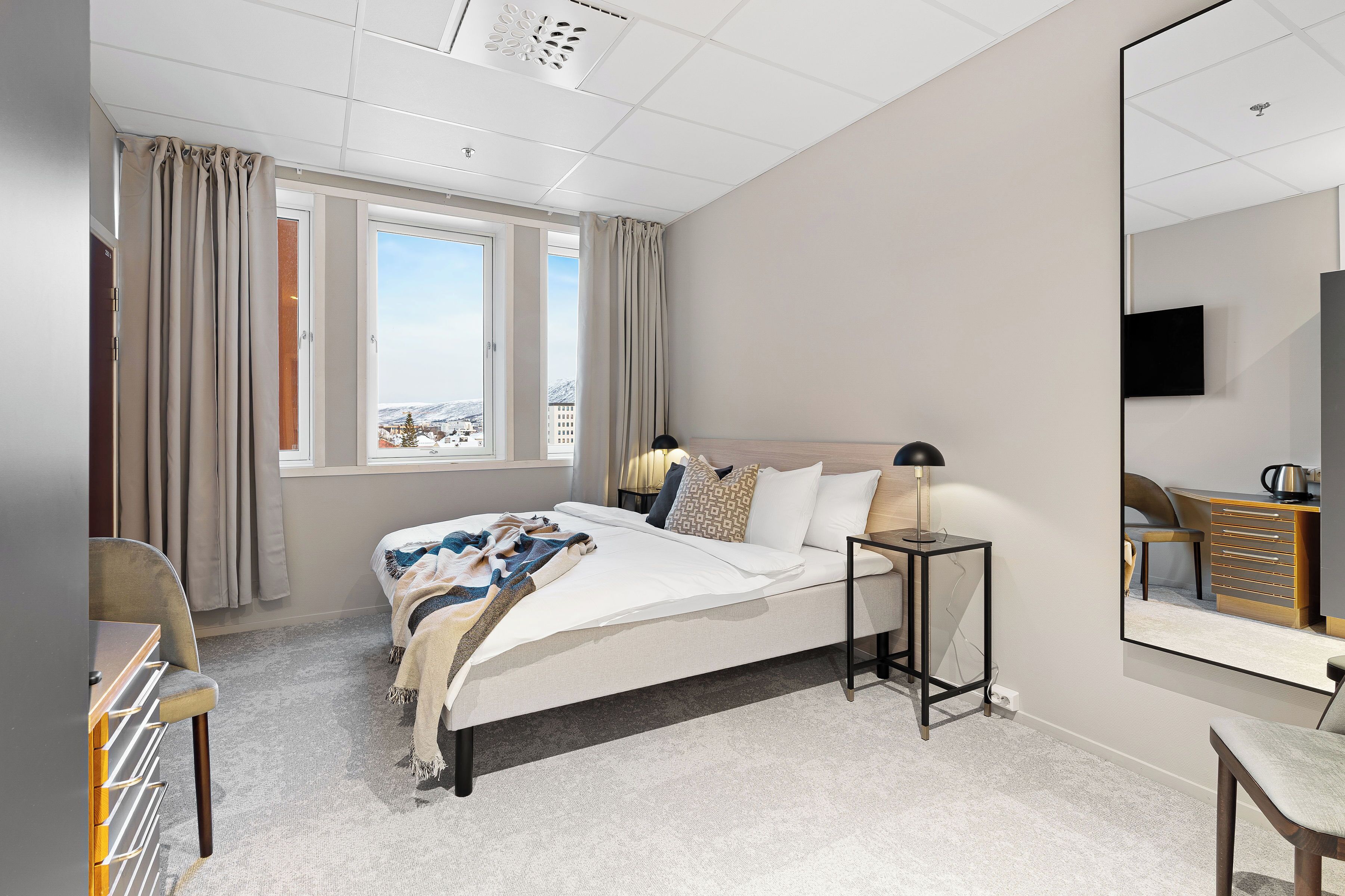 Una camera d'albergo con un letto, comodini, una sedia, una scrivania, uno specchio grande e vista all'esterno.