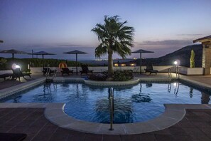 Piscina con palmera, sillas de descanso, sombrillas y vista al mar al atardecer.