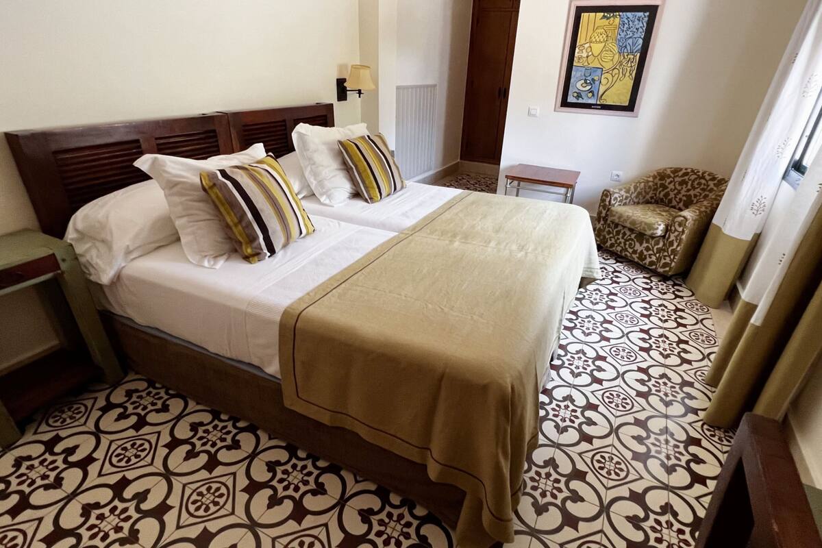 Quarto de hotel com cama de casal, duas mesas de cabeceira, piso estampado, uma cadeira e um quadro na parede.