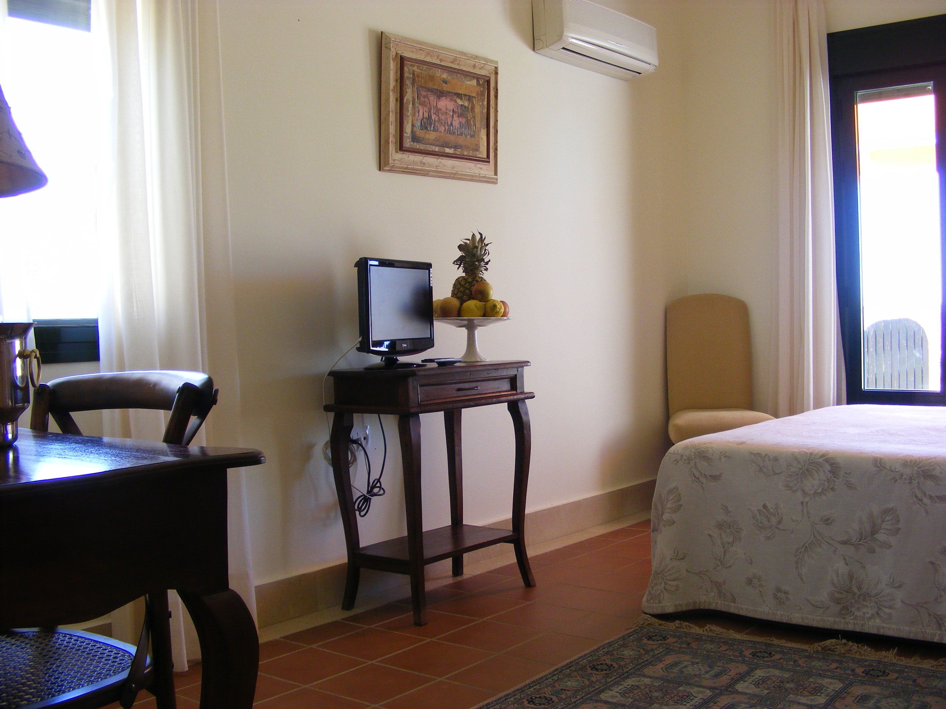 A hotel room with a bed, a desk with a computer, a chair, a small table with a fruit bowl, a framed picture on the wall, and an air conditioner.