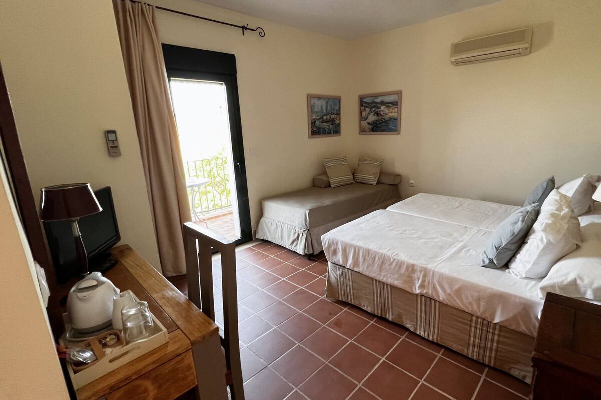 Quarto com cama, luminárias de cabeceira, uma mesinha com abajur, uma cadeira e uma janela com cortinas.