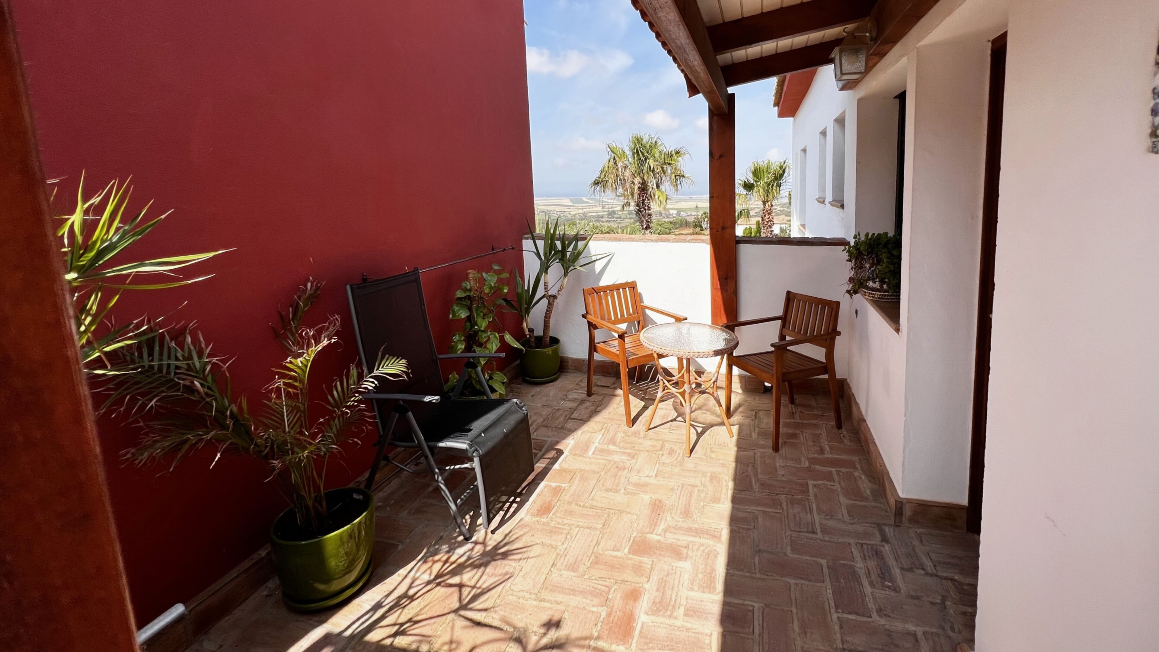 A patio with a red wall, two wooden chairs, a small round table, and potted plants.