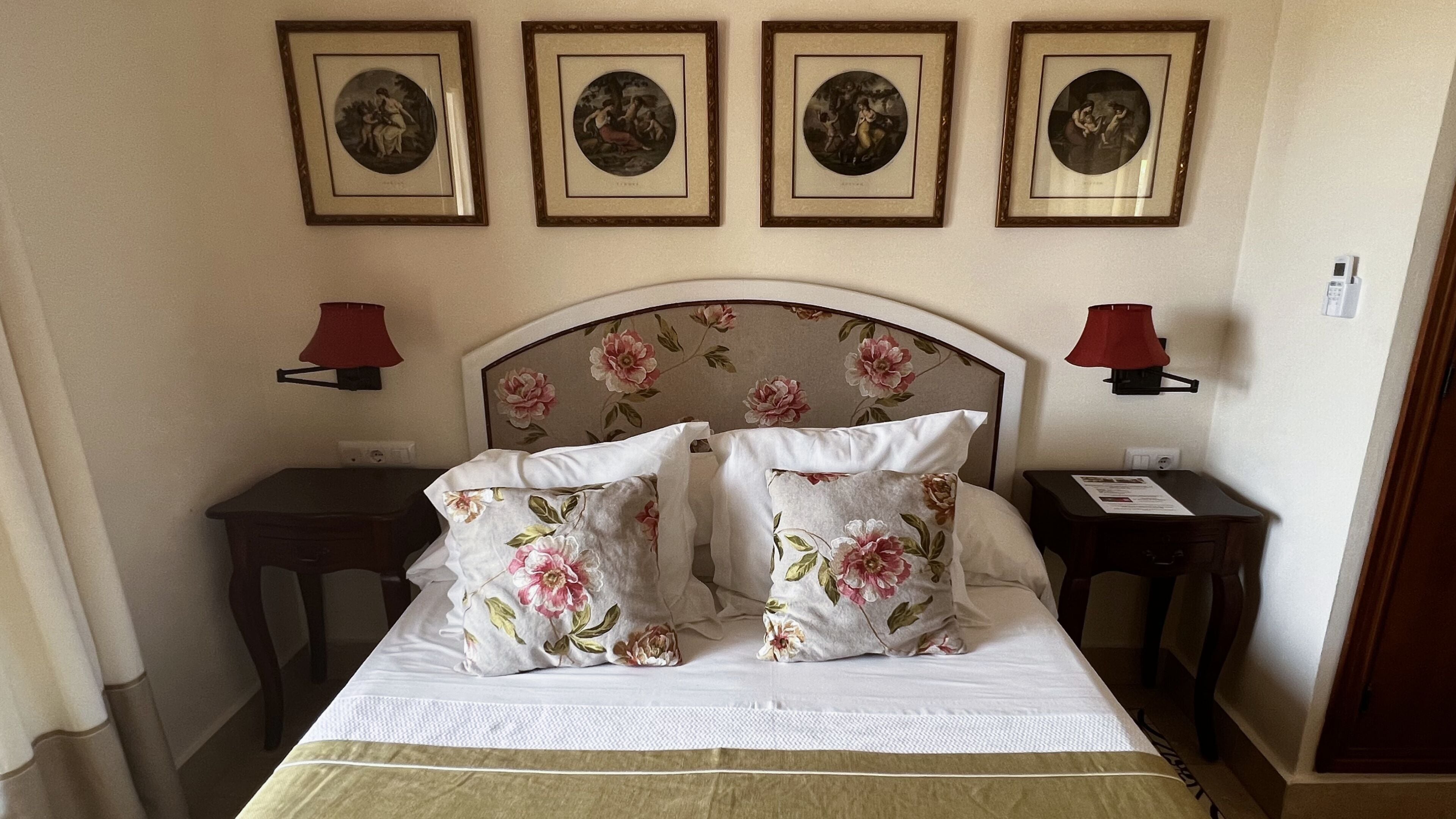 Une chambre d’hôtel avec un lit, une tête de lit à motifs floraux, deux tables de chevet et quatre tableaux encadrés accrochés au mur.