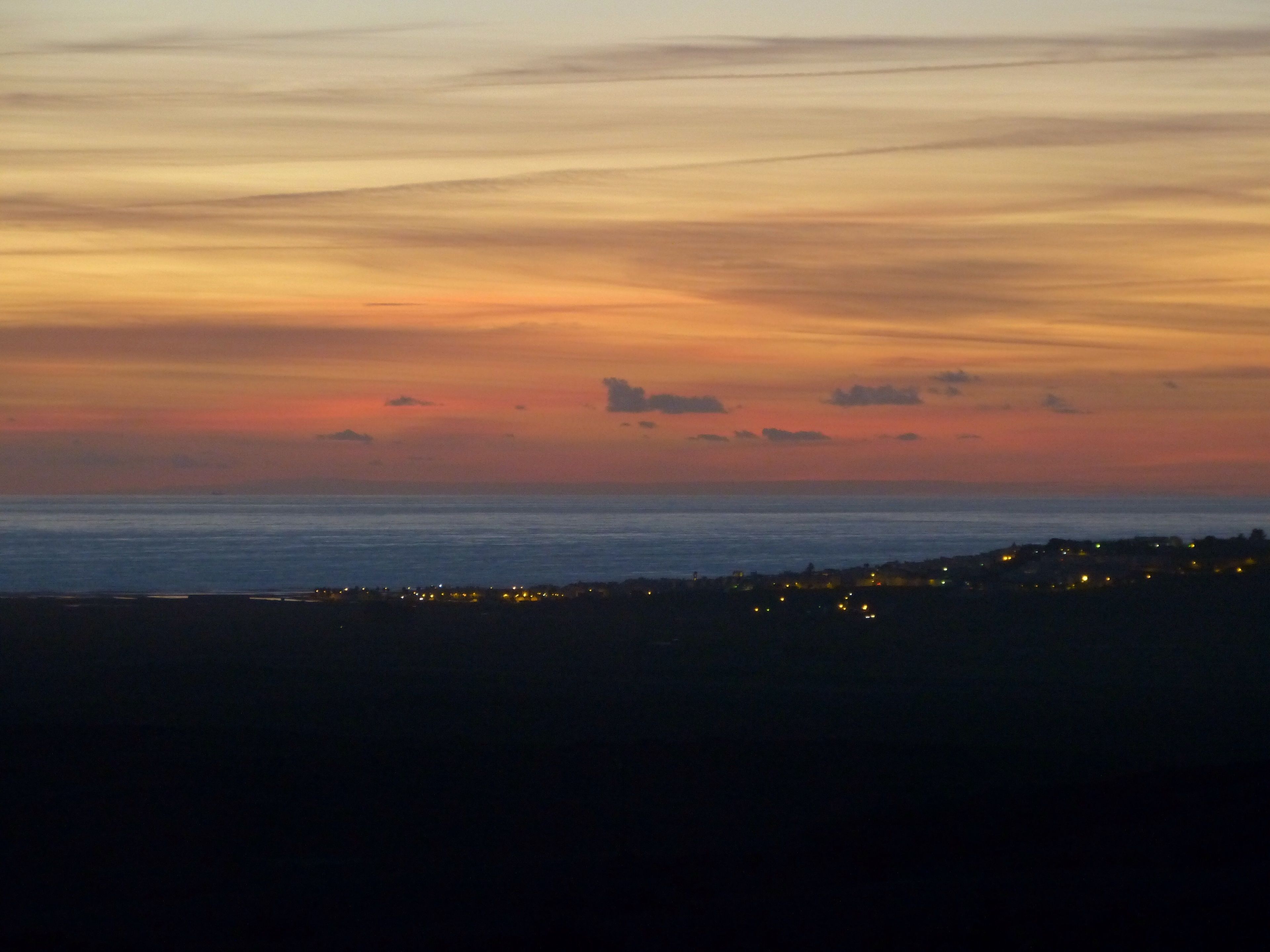 Atardecer costero con vista al océano y una costa distante con luces.