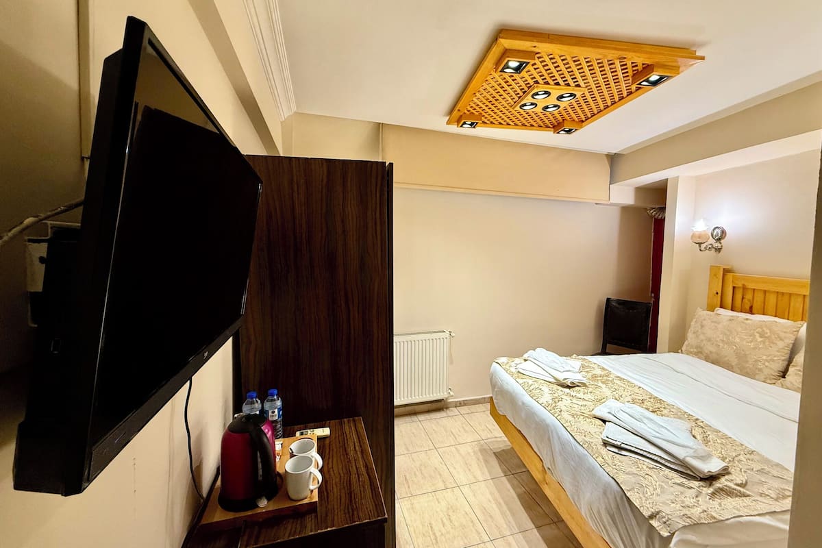 A hotel room with a bed, a TV mounted on the wall, a wooden headboard, a bedside table with a kettle and water bottles, and a ceiling light fixture.
