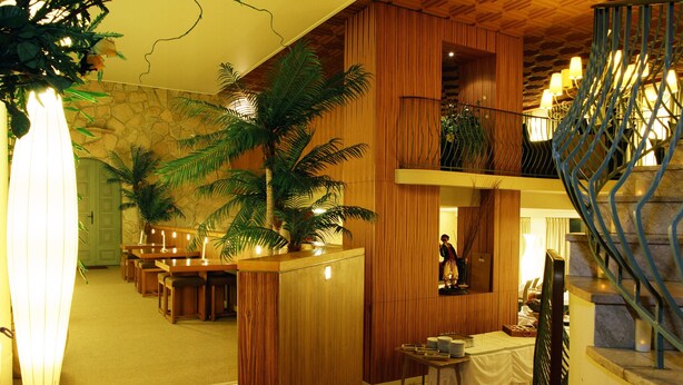 A lobby with a wooden reception desk, a palm tree, a staircase, and a dining area with tables and chairs. - Inter-hotel Au Pavillon Bleu (Ozoir-la-Ferriere)