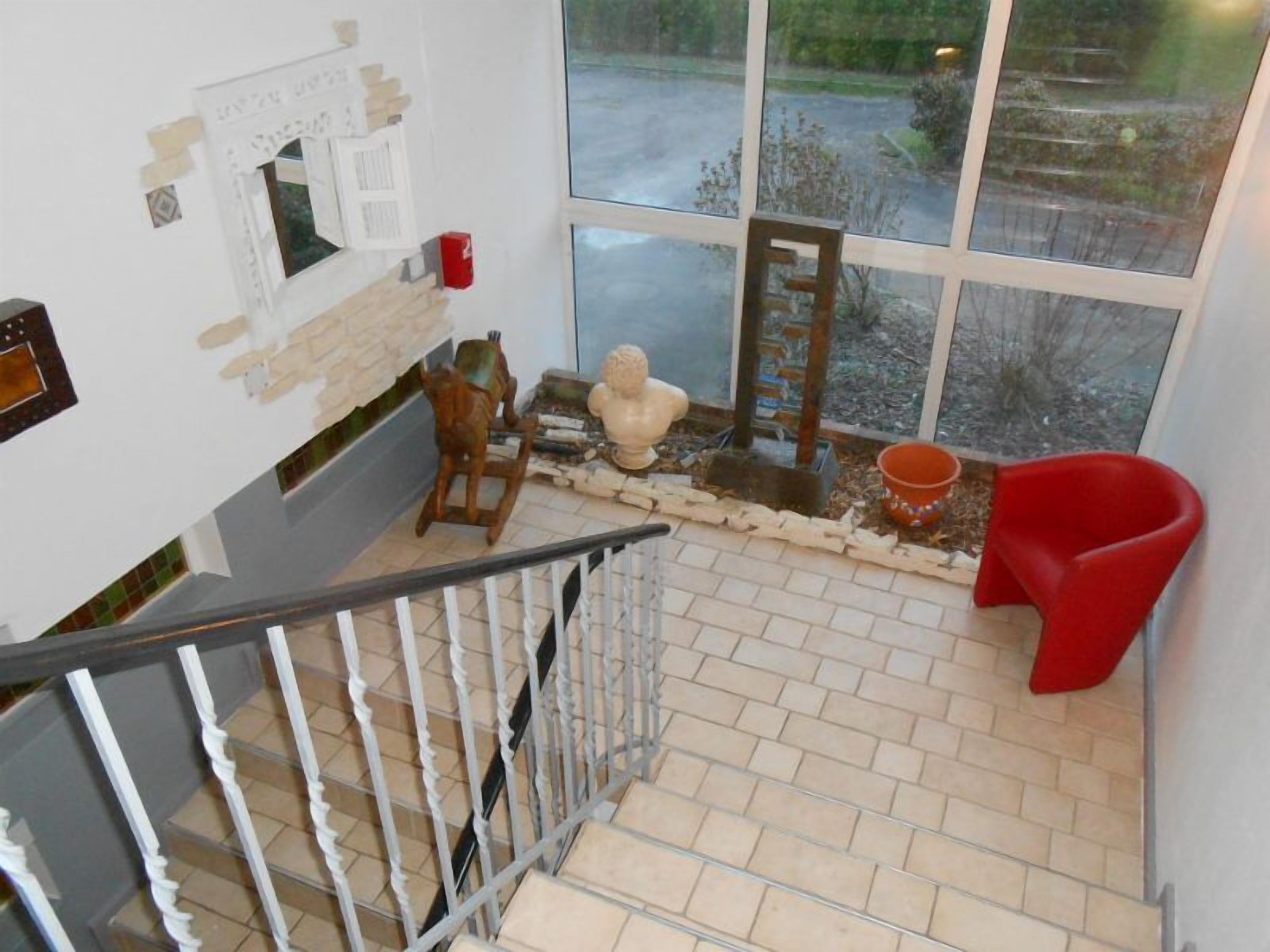 Un escalier avec une rampe en métal mène à une pièce dotée d’une grande fenêtre, d’un fauteuil rouge et d’une petite statue posée sur le rebord de la fenêtre.