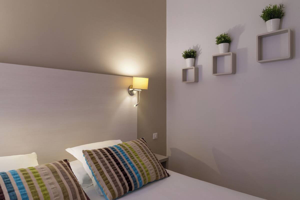 Une chambre d’hôtel bien rangée, avec un lit, des oreillers, une lampe fixée au mur et trois plantes en pot disposées sur le mur.