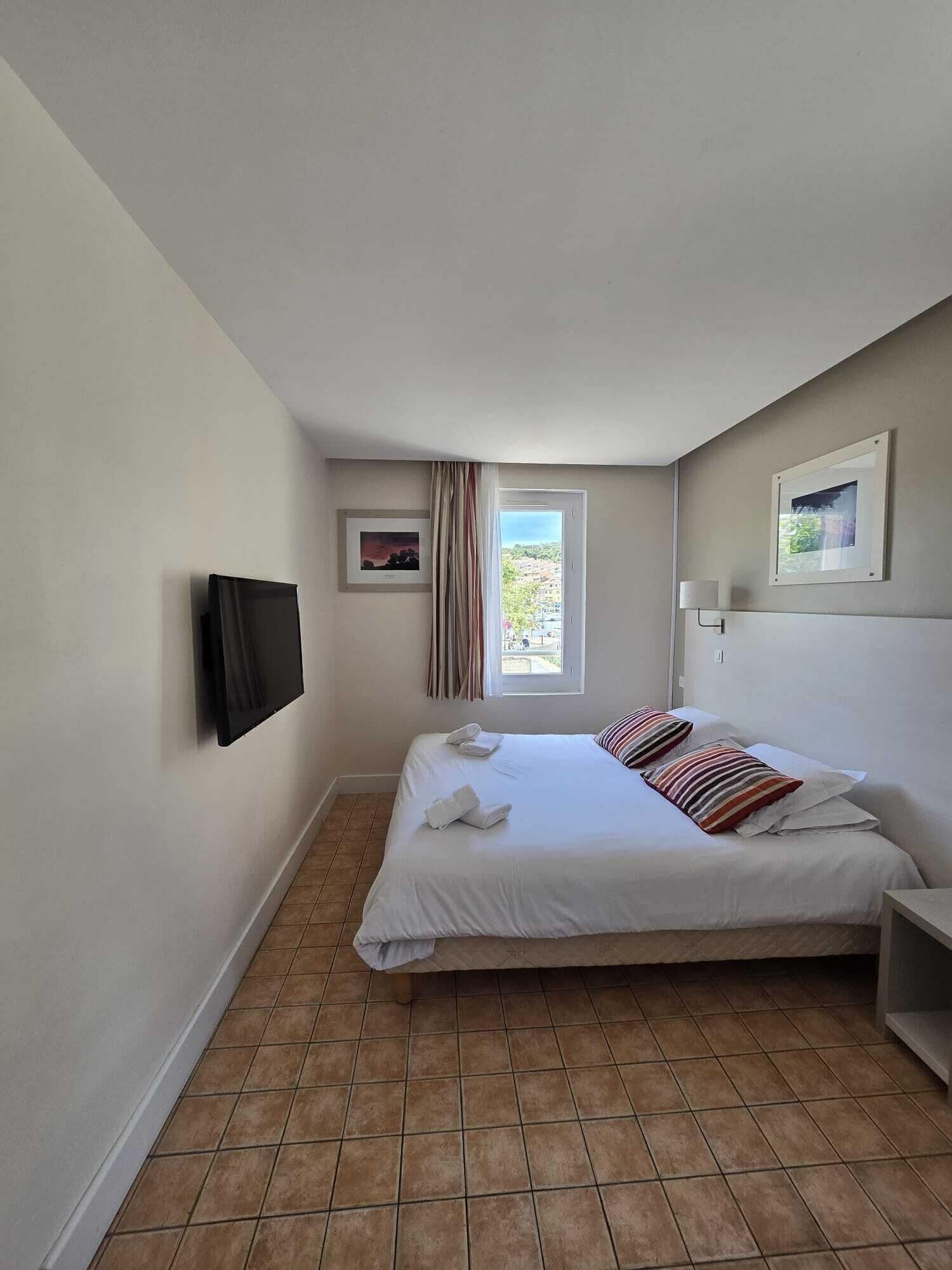Een slaapkamer met een bed, een aan de muur bevestigde televisie, een raam met gordijnen en uitzicht op gebouwen.