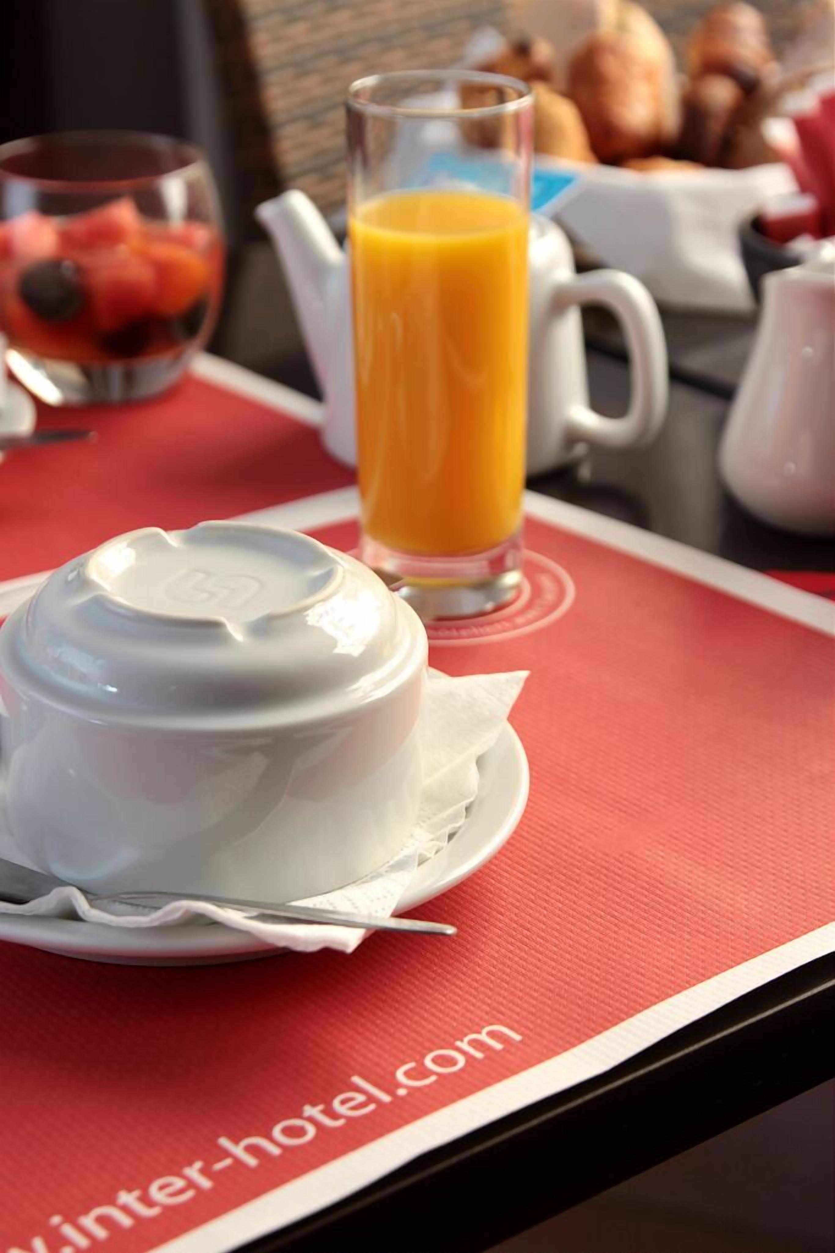 A breakfast setting with a white bowl, a glass of orange juice, and a fruit bowl in the background.