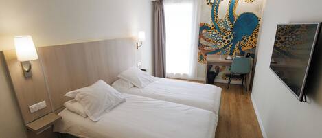A hotel room with two beds, a desk, a chair, a TV, and a colorful tapestry on the wall.