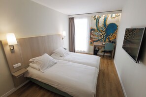 Habitación de hotel con dos camas, un escritorio, una silla, un televisor y un tapiz colorido en la pared.