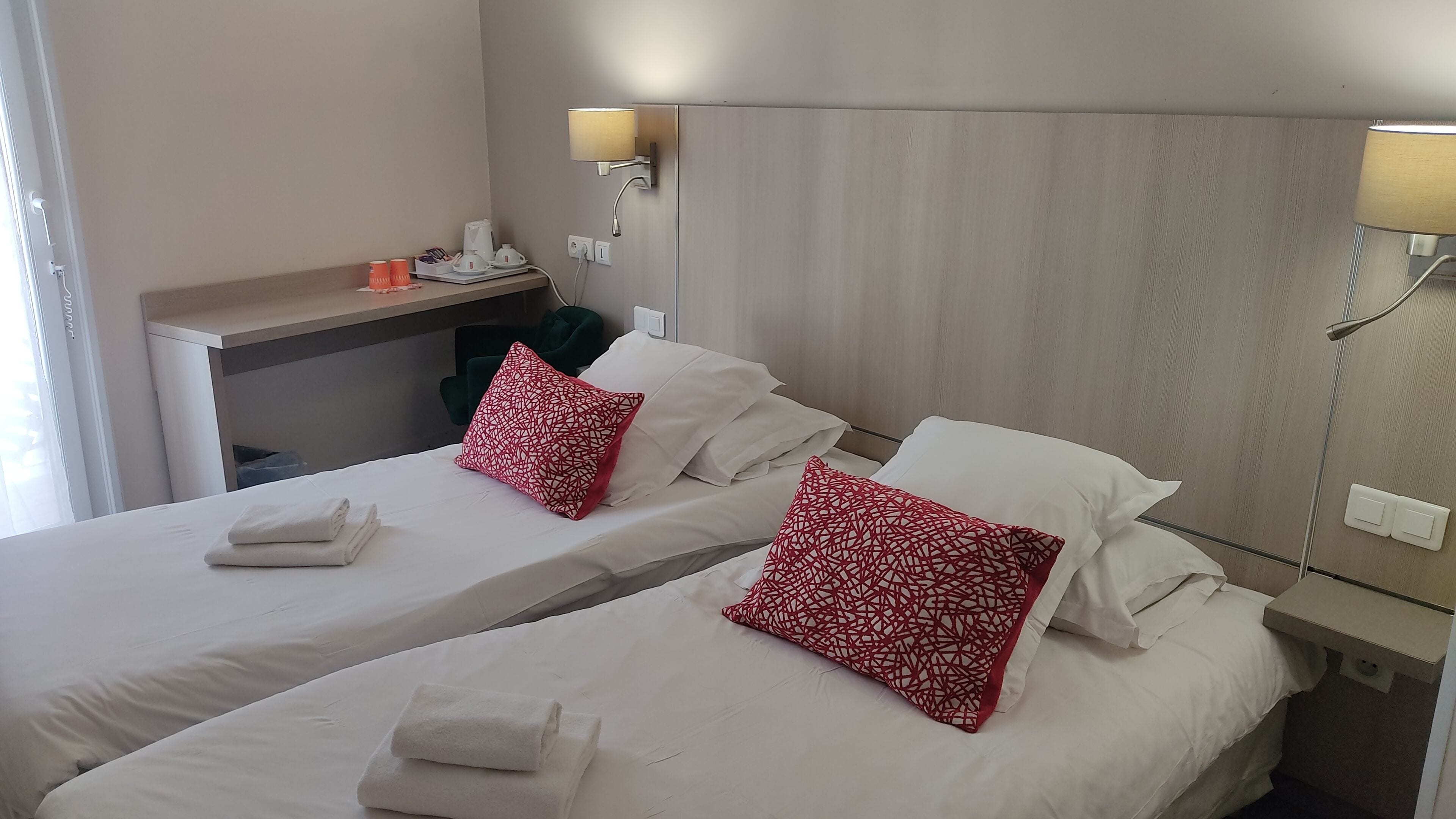 A hotel room with two beds, each with white linens and red patterned pillows. There are two bedside tables with lamps, a small desk with a kettle and cups, and a window with blinds.