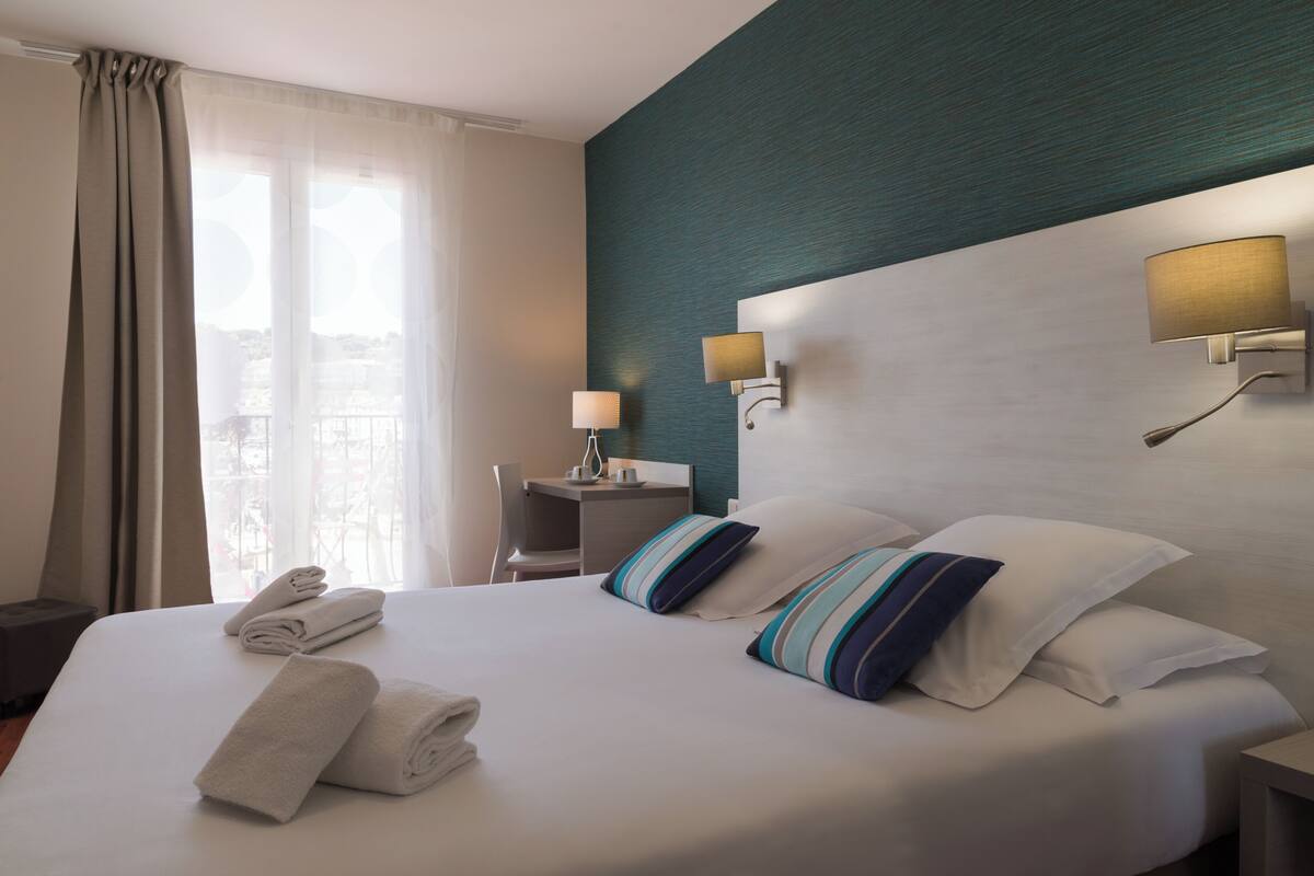 Une chambre d’hôtel avec un lit parfaitement fait, deux lampes de chevet, une petite table avec une lampe et une vue sur la ville grâce à de grandes fenêtres.