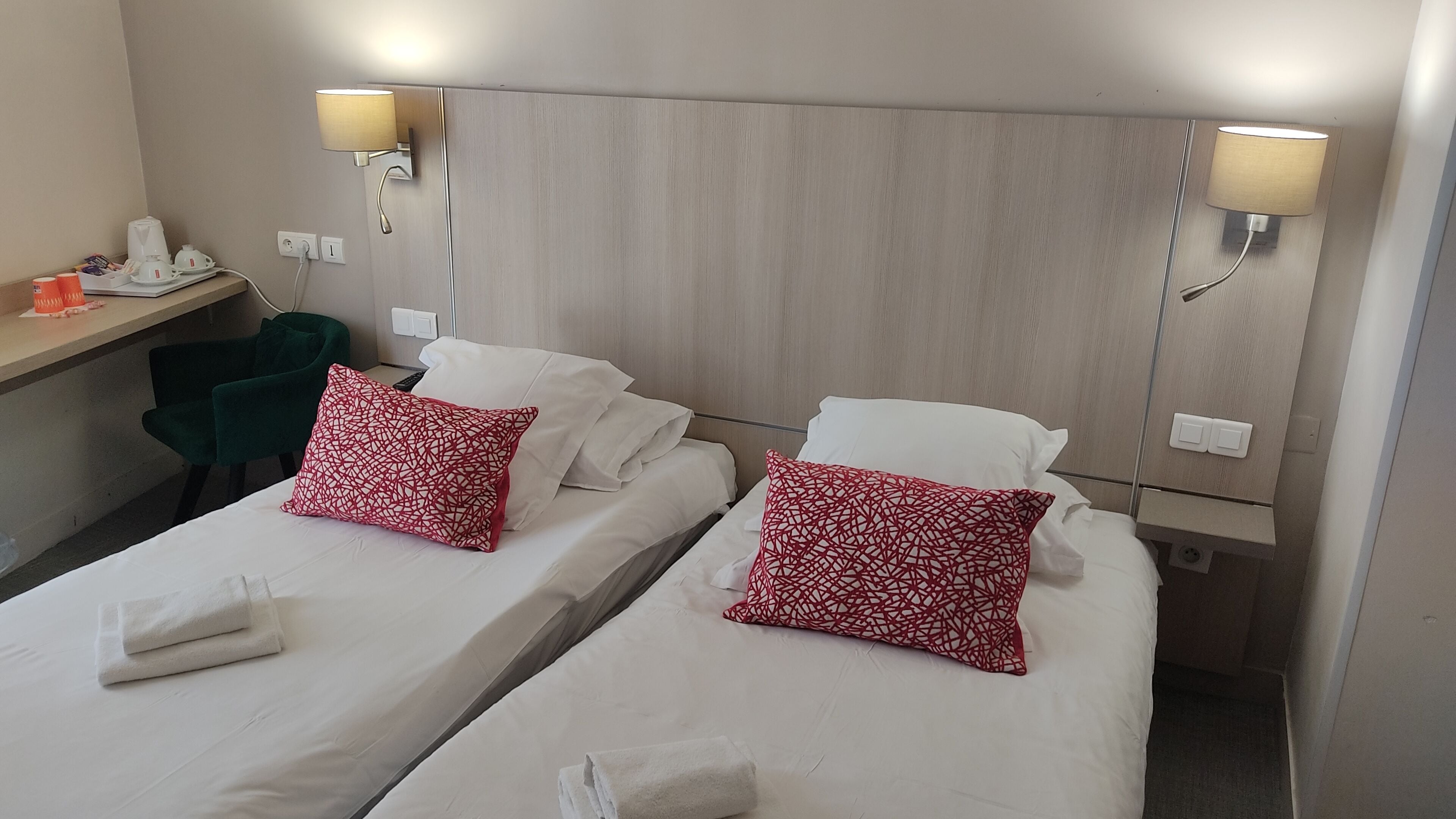 A hotel room with two beds, each with white linens and red patterned pillows. There are bedside lamps, a desk with a phone and a coffee maker, and a green chair.