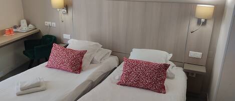 A hotel room with two beds, each with white linens and red patterned pillows. There are bedside lamps, a desk with a phone and a coffee maker, and a green chair.