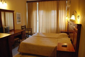 A hotel room with two beds, a desk with a television, a chair, and a nightstand with a lamp. - Galini Hotel (Aegina)