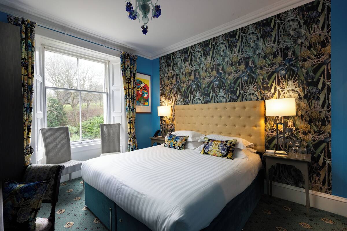 A bedroom with a large bed, two bedside tables, a window with curtains, and a wall with a floral wallpaper.